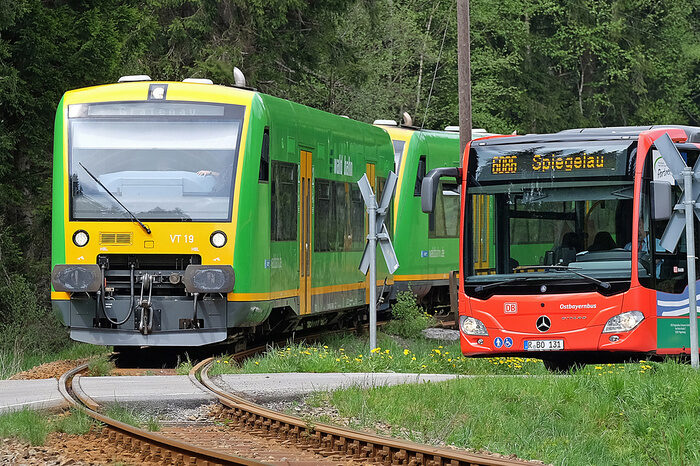 Die Waldbahn steht an einem Bahn&uuml;bergang neben einem Igelbus im Bayerischen Wald.